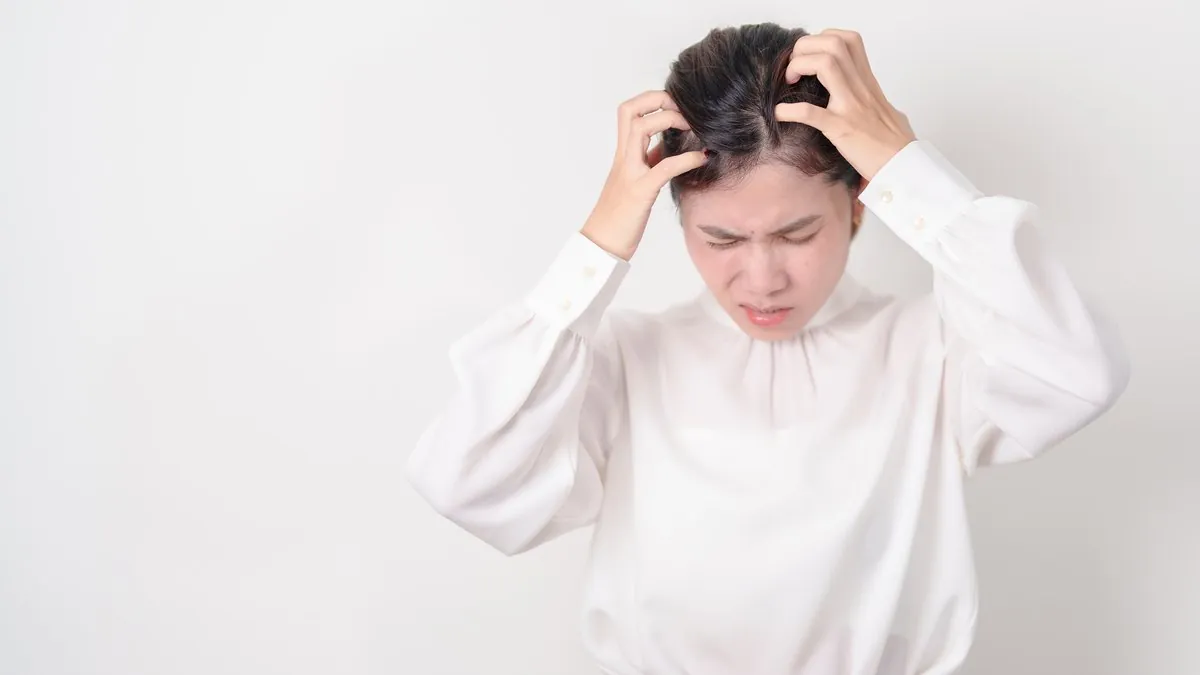 Femeie în bluză albă își scarpină scalpul cu ambele mâini, cu o expresie de disconfort – dermatita seboreică