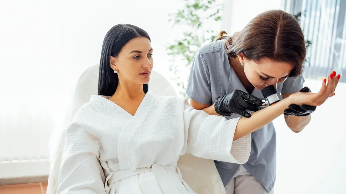 Dermatolog examinează cu dermatoscopul pielea antebrațului unei paciente în halat alb – depistarea alunițelor atipice